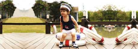 Toddler in navy tank top and shorts with sailor hat sitting on dock by life buoys outdoors