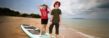 Kids in summer outfits and sunglasses posing on a beach by a surfboard