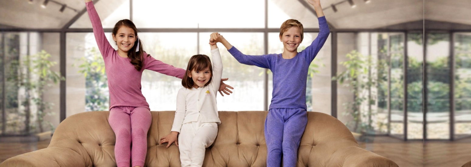 Three kids in colorful winter thermal innerwear smiling on a sofa in a bright living room