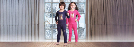 Two children in colorful winter thermal innerwear sets standing indoors with snowy window backdrop