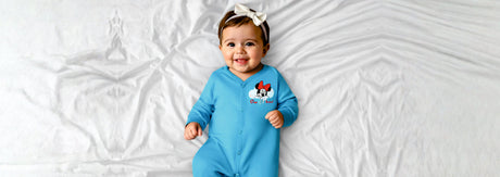 Smiling baby in blue cartoon-print romper and bow headband lying on white bed